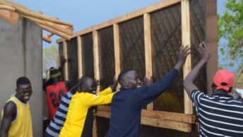 Photo of 4 men holding up a wooden wall and one man behind them. All are laughing and appear to be jovial